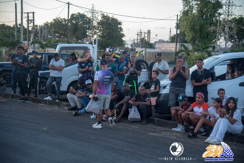 A group of people standing and sitting beside parked vehicles near the course.