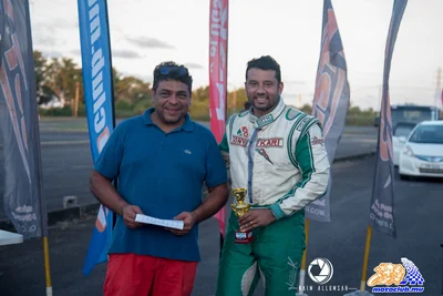 Bill Ghanty on the left, in a blue polo and red shorts, beside a racer holding a trophy.